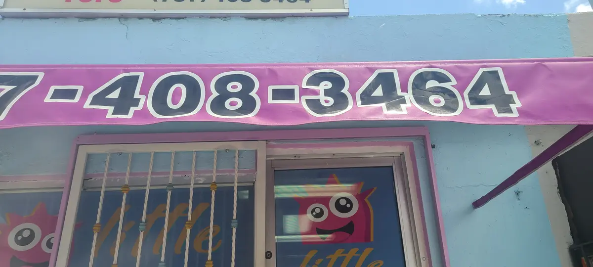 Fachada de Little Cuts: pared azul cielo, toldo rosado con el teléfono y la mascota de la barbería en la puerta.