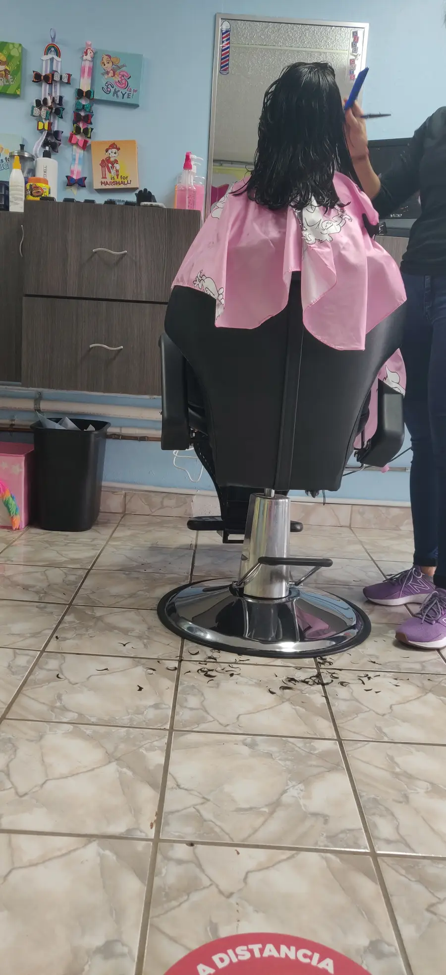 Una niña de espaldas con capa rosada en la silla de la barbería; en la pared, cuadros de personajes infantiles y lazos de colores.